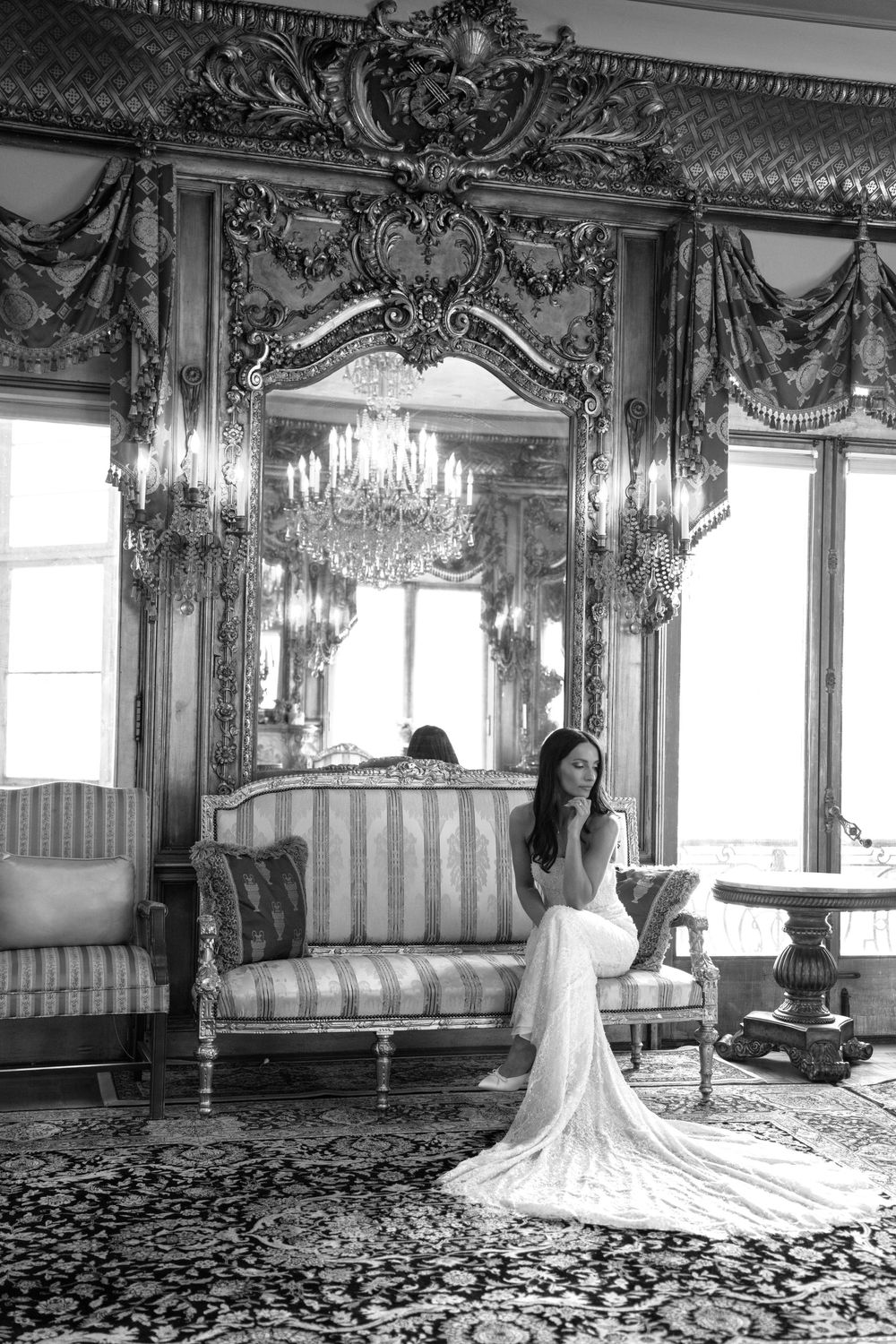 Bride in white gown sits on striped sofa in ornate gilded room with chandelier, black and white photo.