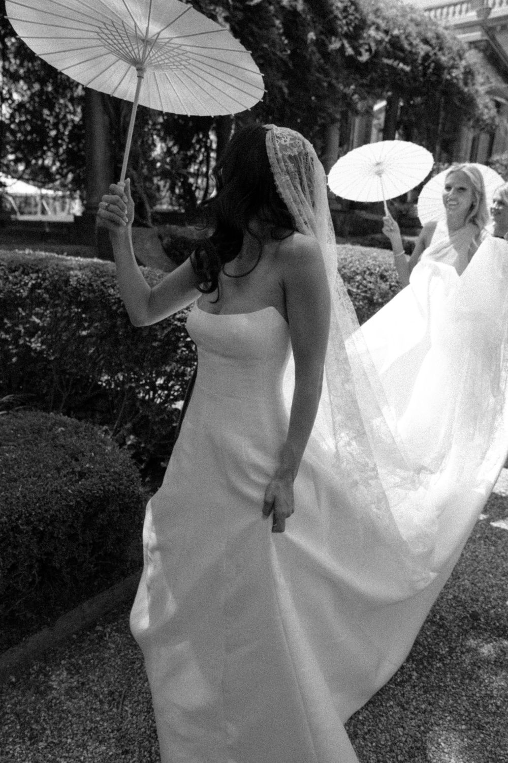 Two brides in white gowns holding parasols outdoors in a black and white wedding photo.