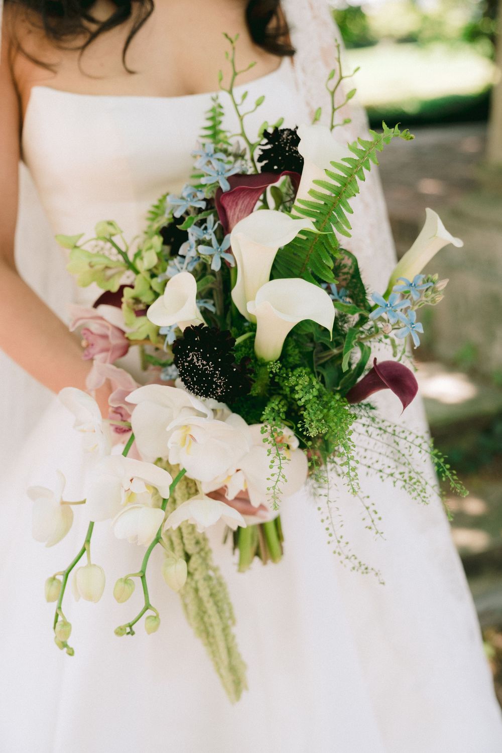 Bride holding a lush wedding bouquet with white calla lilies, orchids, ferns, and dark purple blooms.