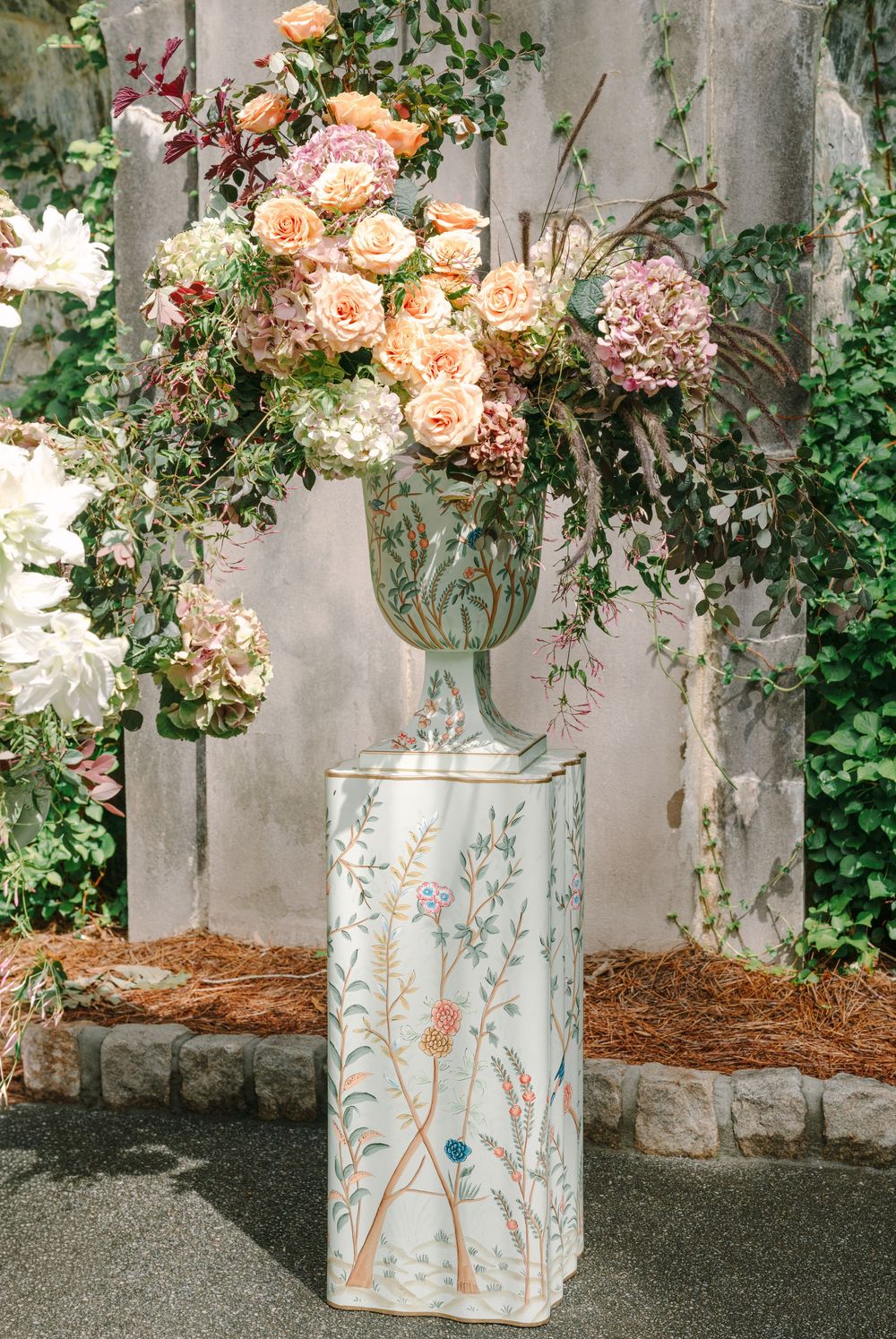 Elegant floral arrangement with peach roses and hydrangeas in a painted urn on a decorative pedestal outdoors.