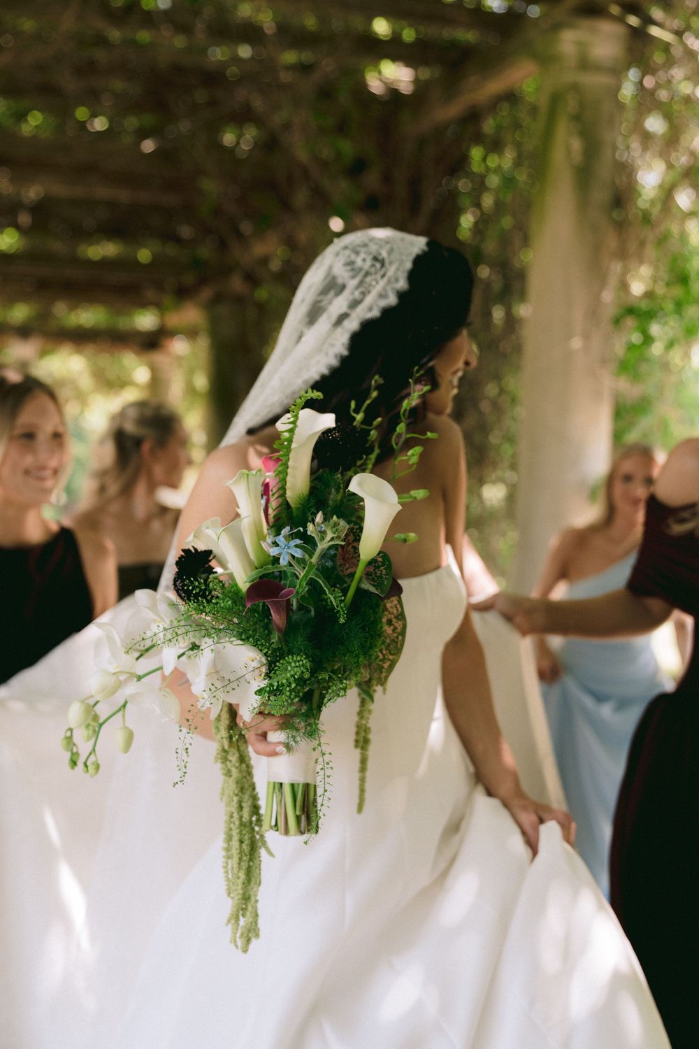 Bride in white gown and lace veil holds lush bouquet of calla lilies and greenery, surrounded by bridesmaids in garden.