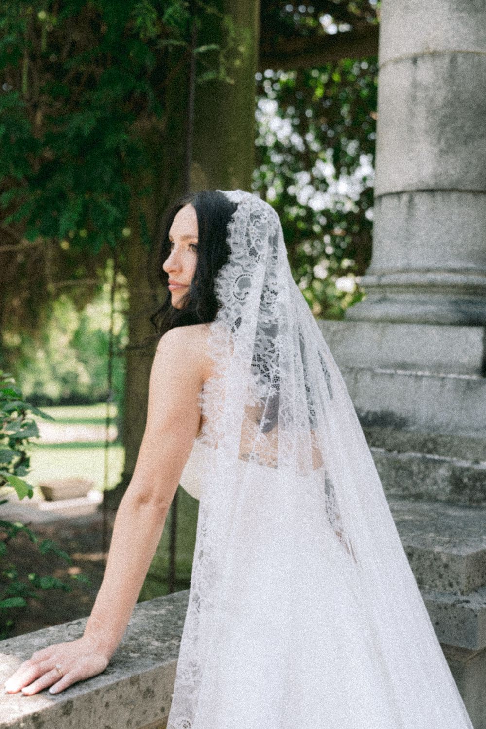 Bride in lace veil and strapless gown poses by stone column in lush garden setting.