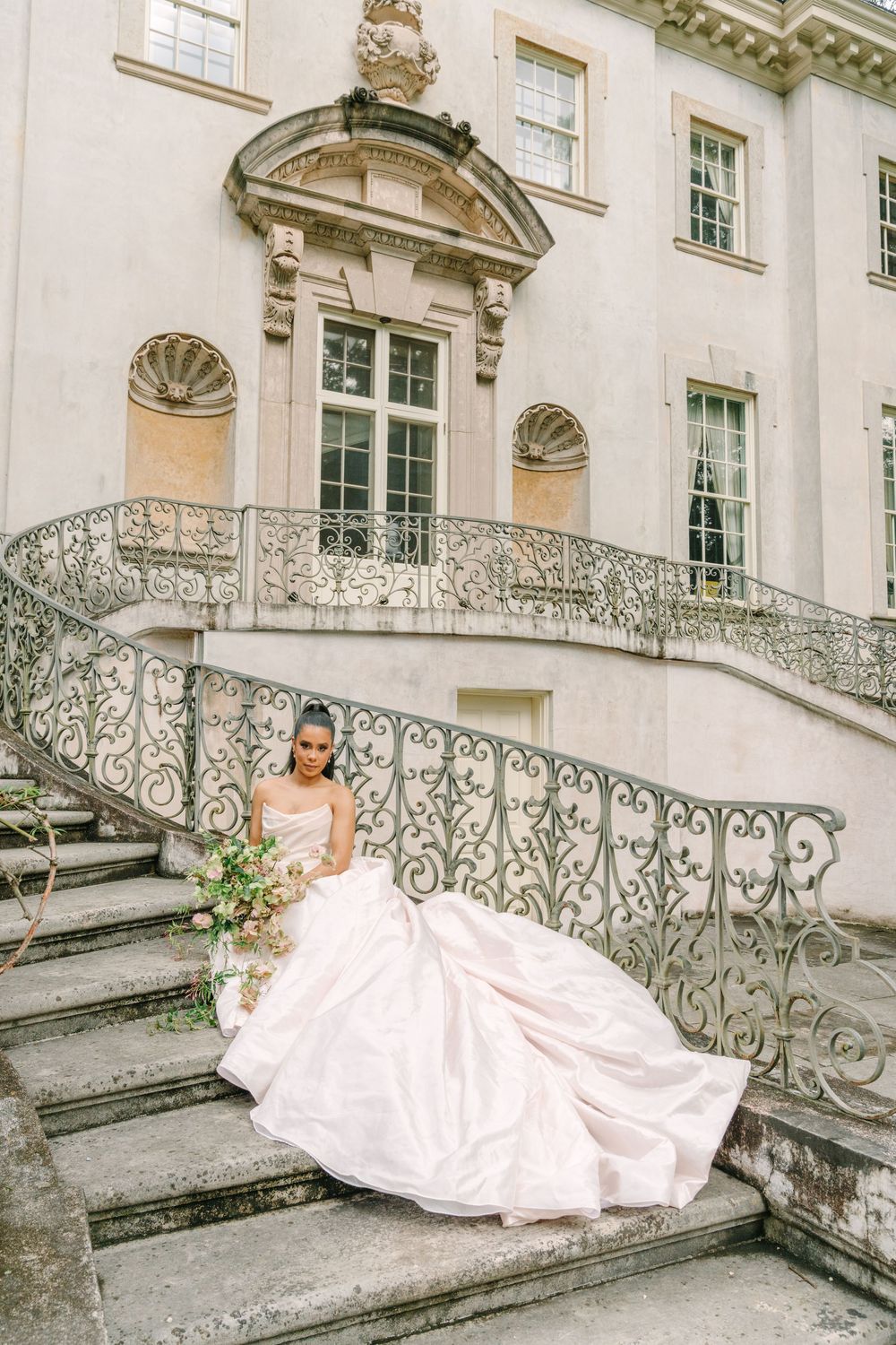 Bride in a flowing white gown sits on ornate stone staircase of elegant mansion holding a bouquet.