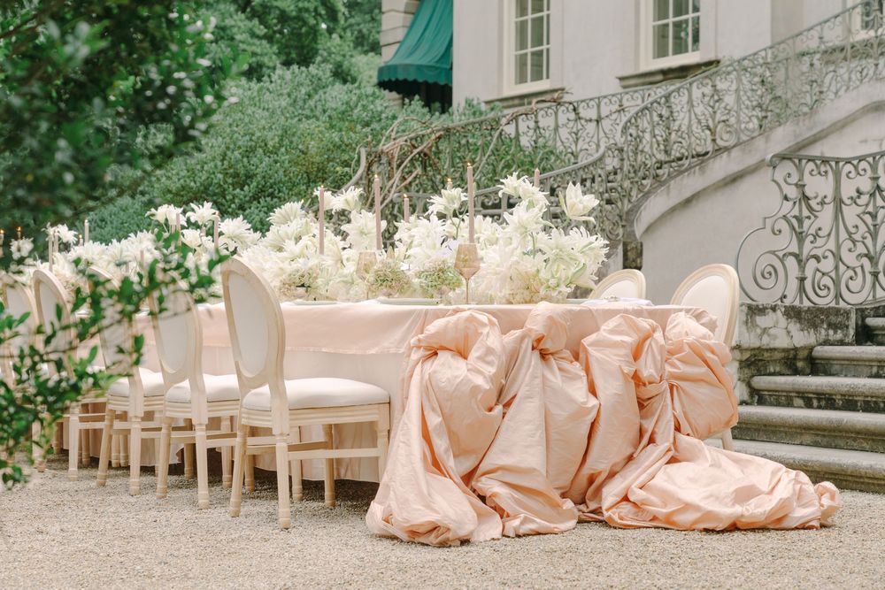Elegant outdoor wedding reception table with blush pink linen, white floral centerpieces, and ornate chairs near a grand staircase.