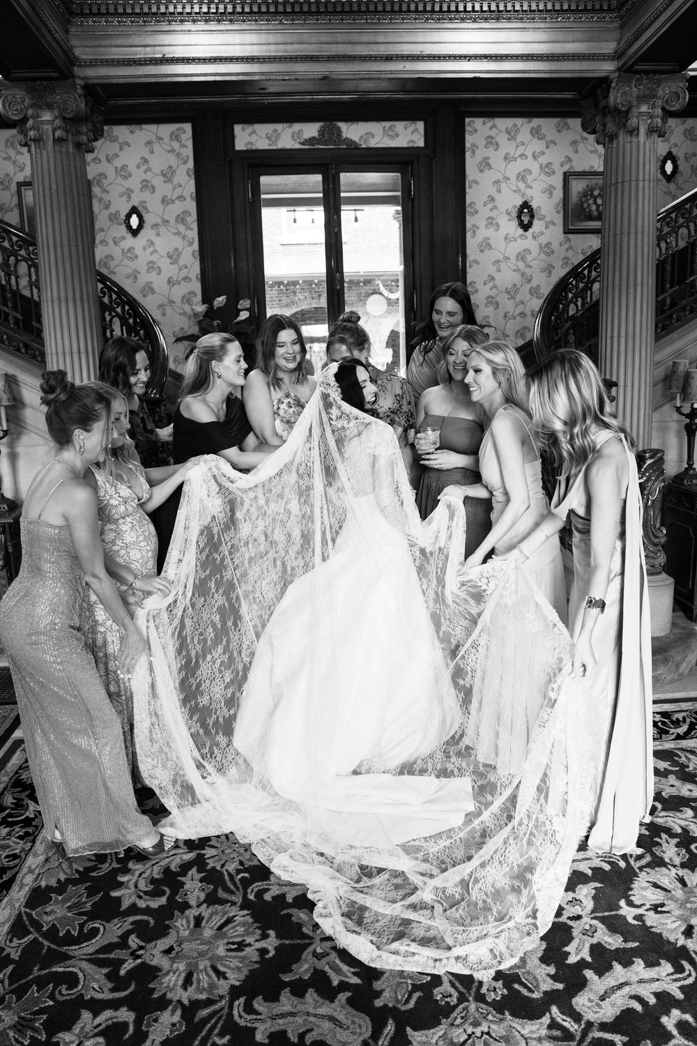 Bridesmaids surround bride holding lace veil train in elegant staircase foyer, black and white wedding photo.