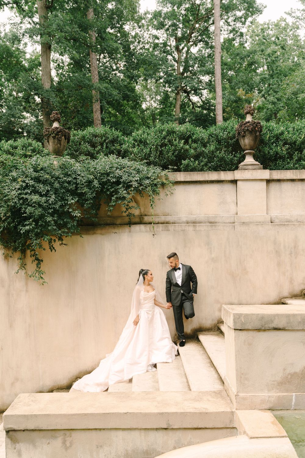 Bride and groom hold hands on stone steps surrounded by lush greenery at an elegant outdoor wedding venue.