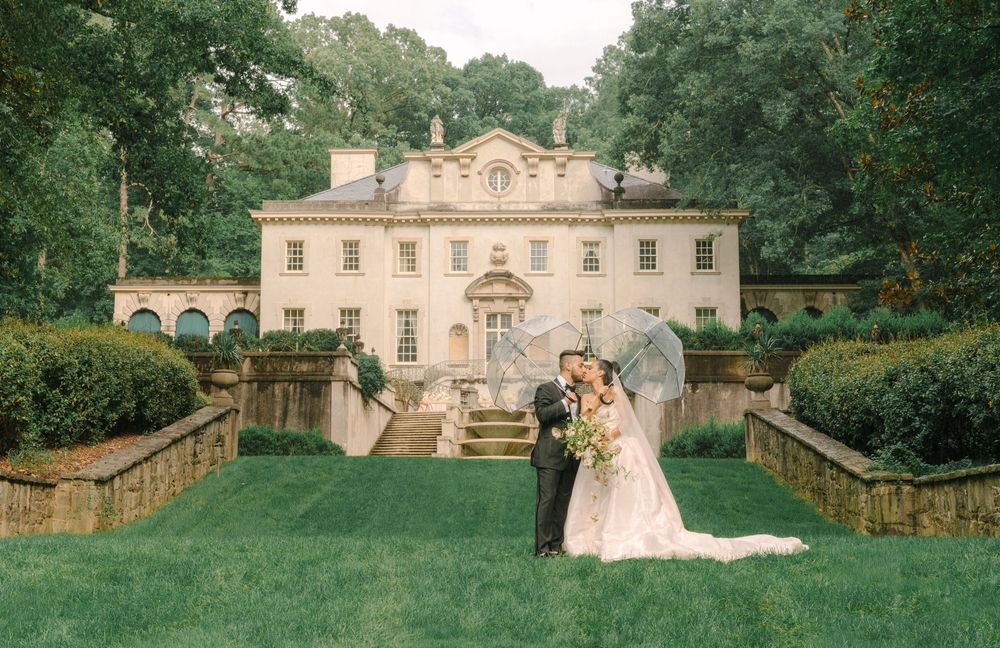 Bride and groom share a romantic kiss outside an elegant Georgian mansion surrounded by lush gardens.