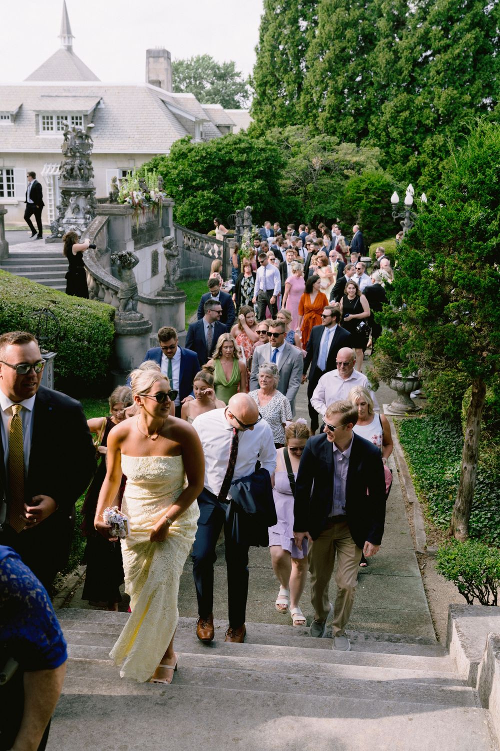 Wedding guests walking down ornate stone steps at elegant estate venue surrounded by lush greenery.