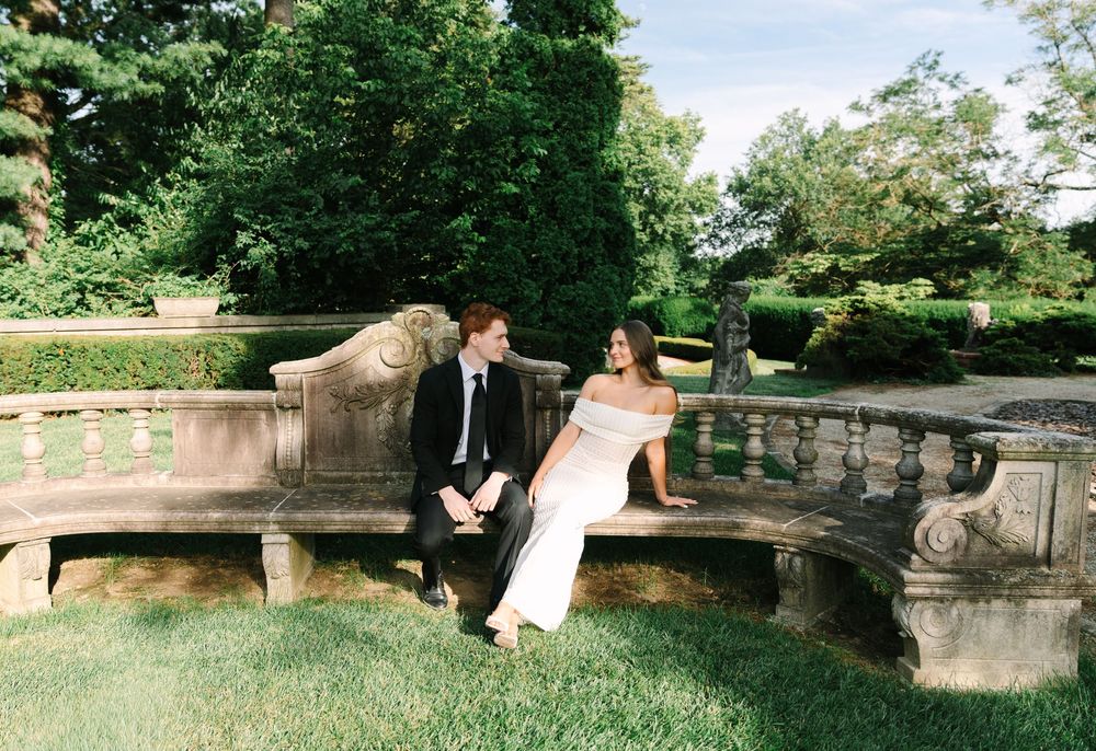 Elegant couple in formal attire sitting on a stone bench surrounded by lush greenery in a garden setting.