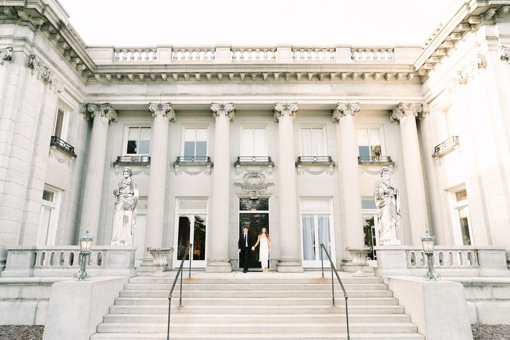 Couple stands at entrance of grand neoclassical building with white columns and ornate statues on wedding day.