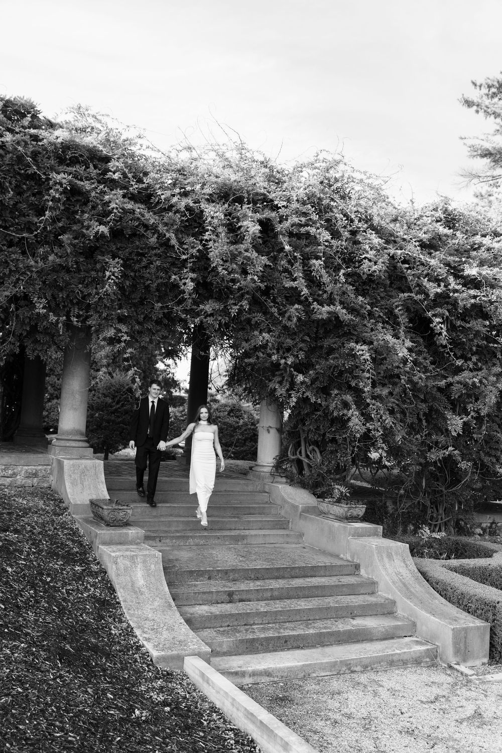 Black and white photo of a bride and groom holding hands walking down stone steps in a lush garden.