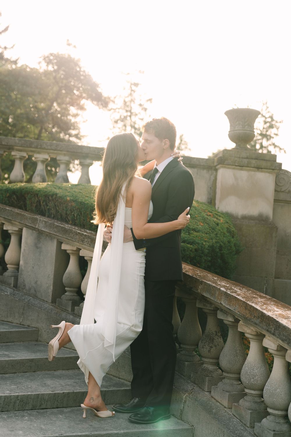 Bride and groom share a romantic moment on stone steps at golden hour, surrounded by lush greenery.