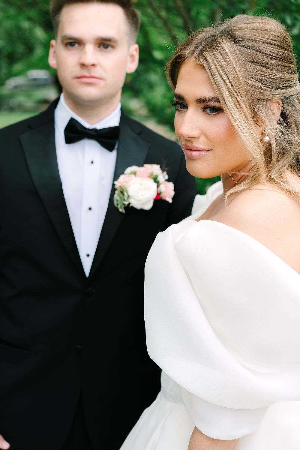 Bride in off-shoulder white gown and groom in black tuxedo with boutonniere pose outdoors at wedding.
