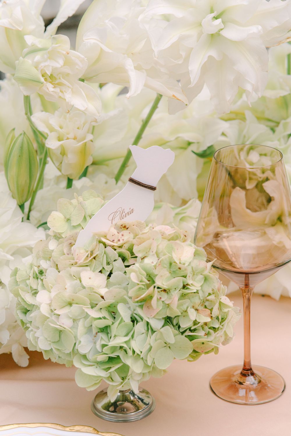 Elegant wedding table with white hydrangeas, orchids, place card, and rose gold wine glass on cream tablecloth.