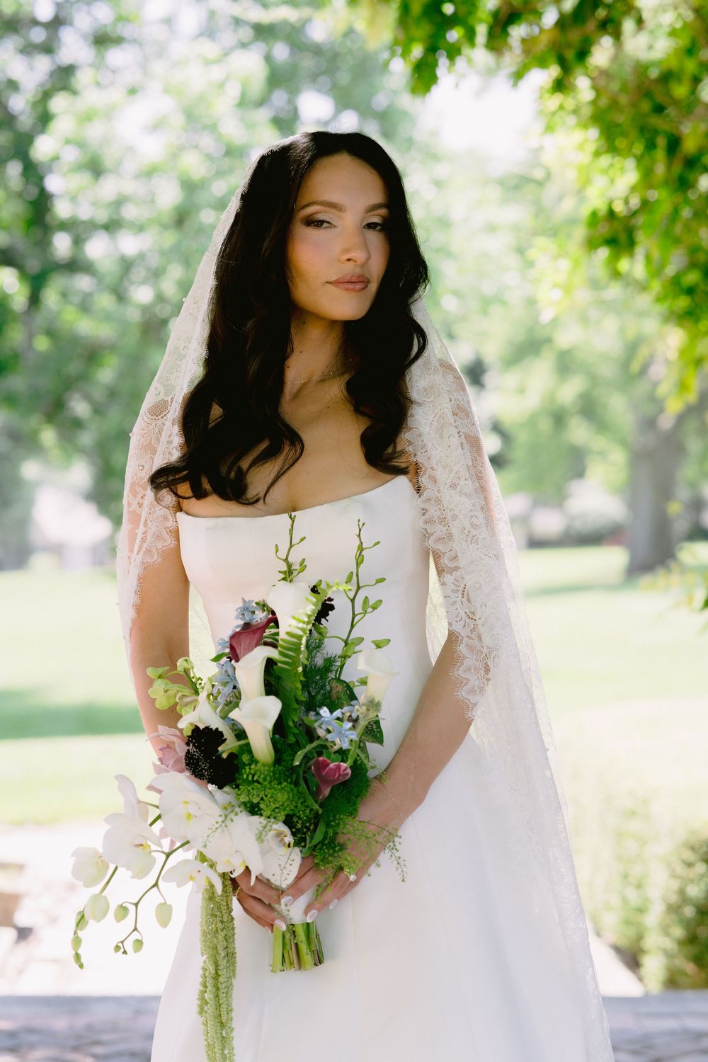 Bride in strapless white gown with lace veil holding lush floral bouquet outdoors.