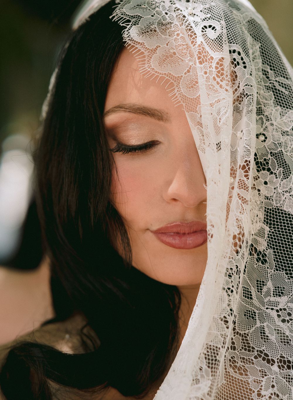 Bride wearing an elegant white lace veil with floral pattern, dark hair, natural makeup, eyes closed.