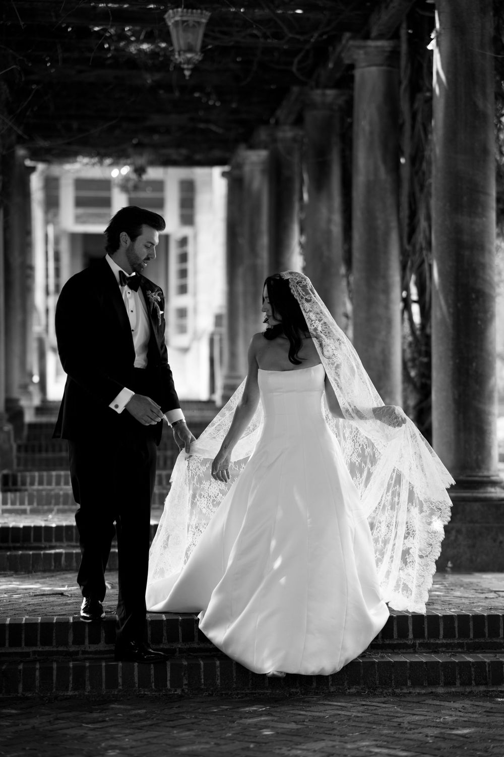 Black and white photo of bride and groom holding hands on steps at elegant columned venue.