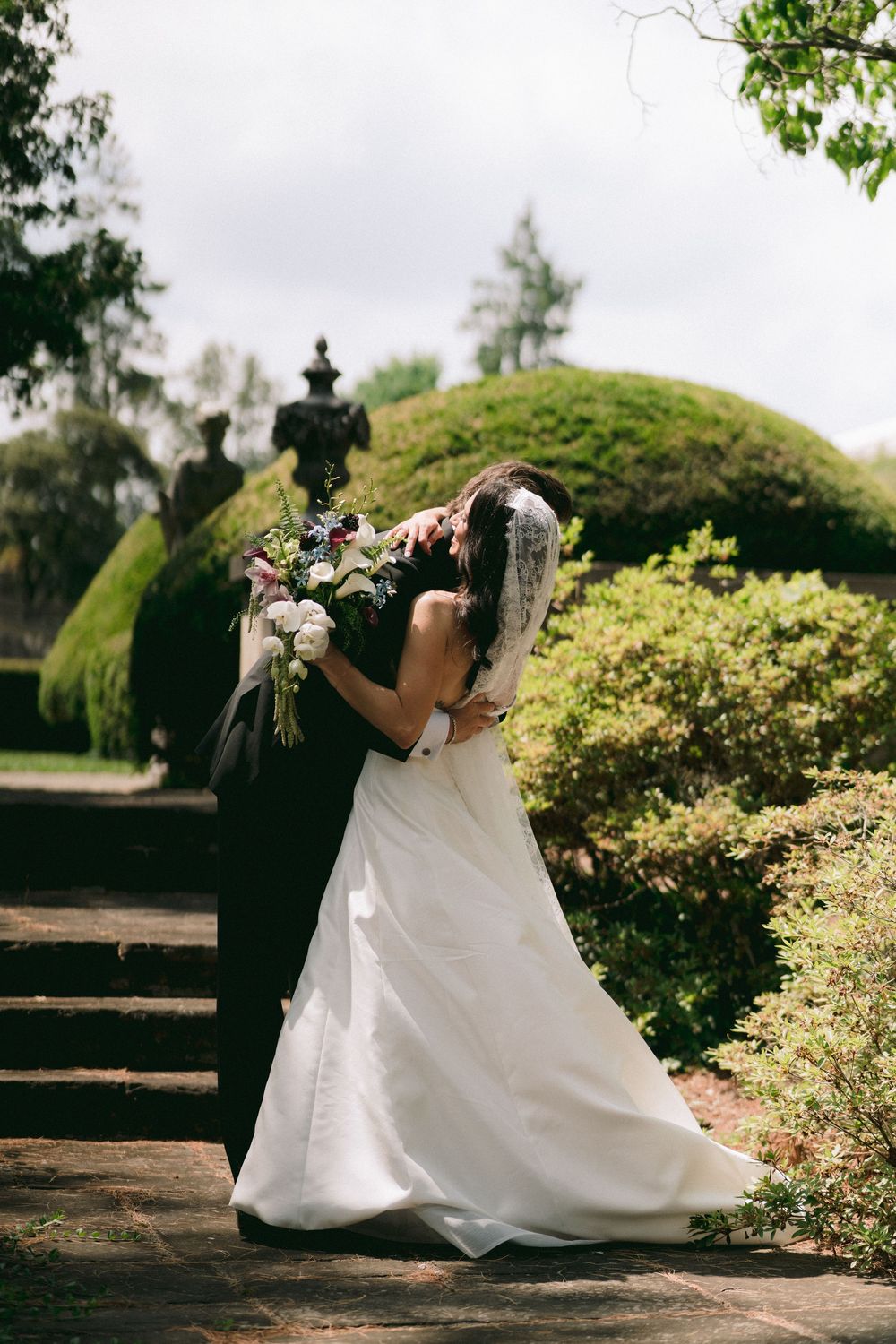 Bride and groom share a romantic embrace in a lush garden, bride holding a beautiful floral bouquet.
