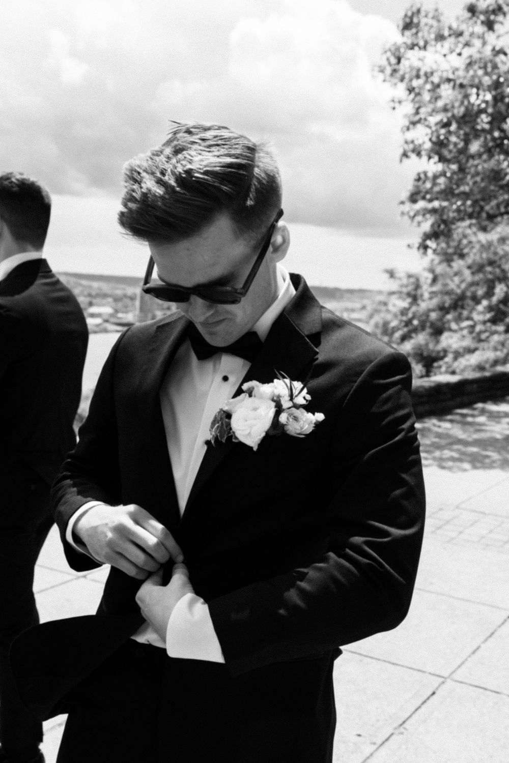 Black and white photo of a groom in a tuxedo with boutonniere and sunglasses adjusting his cufflinks outdoors.