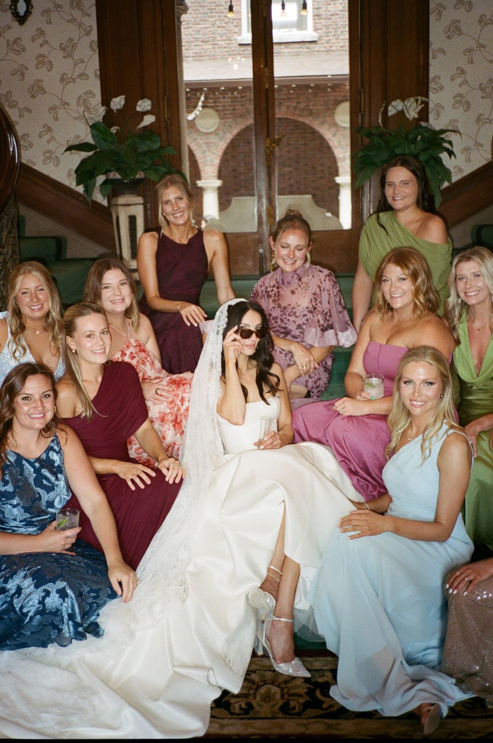 Bride in white gown and veil poses with bridesmaids in colorful dresses on elegant staircase.