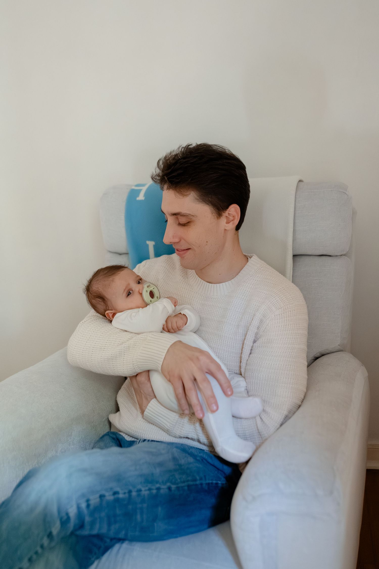 Young father in white sweater sits in gray chair, lovingly holding newborn baby with pacifier in cozy nursery.