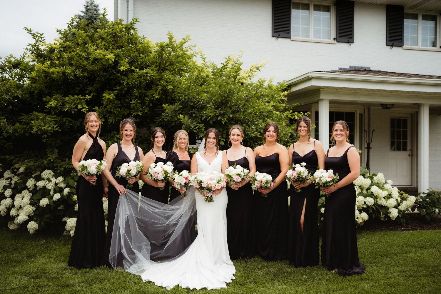 Bride in white gown with bridesmaids in black dresses holding white bouquets, posing outdoors near white hydrangeas.