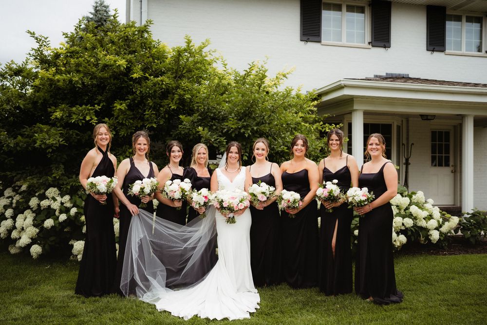 Bride in white gown with bridesmaids in black dresses holding white bouquets, posing outdoors near white hydrangeas.