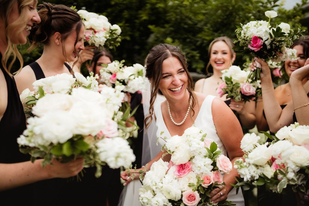 Bride smiling with bridesmaids holding white and pink floral bouquets at an outdoor wedding celebration.