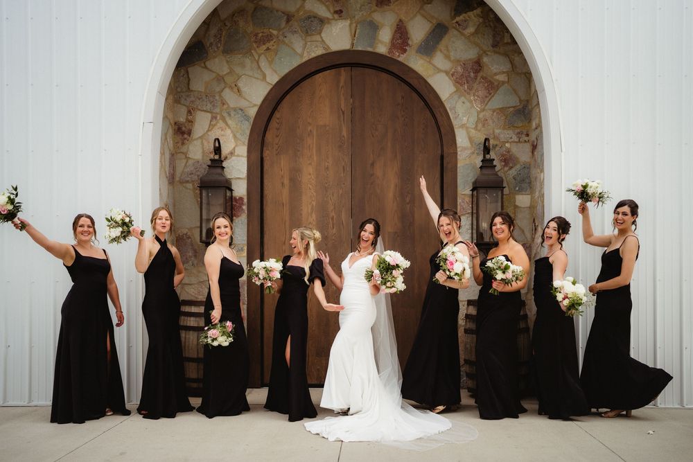 Bridesmaids in black gowns celebrate with bride in white dress before rustic wooden arched door.