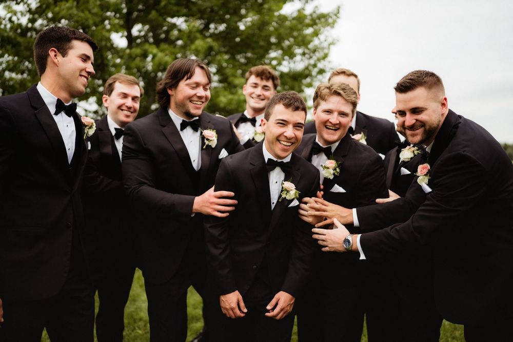 Groomsmen in black tuxedos with boutonnieres laughing together outdoors at a wedding celebration.