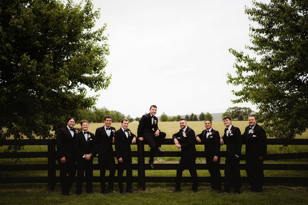 Groomsmen in black tuxedos posing by a wooden fence with lush green trees and fields in the background.
