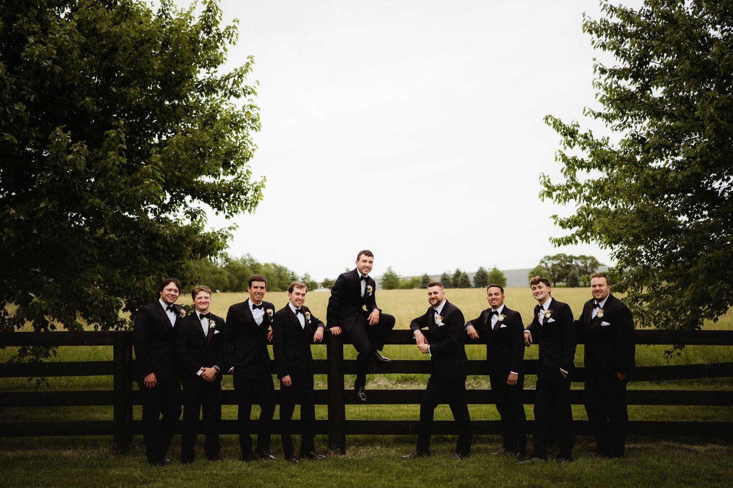 Groomsmen in black tuxedos posing by a wooden fence with lush green trees and fields in the background.