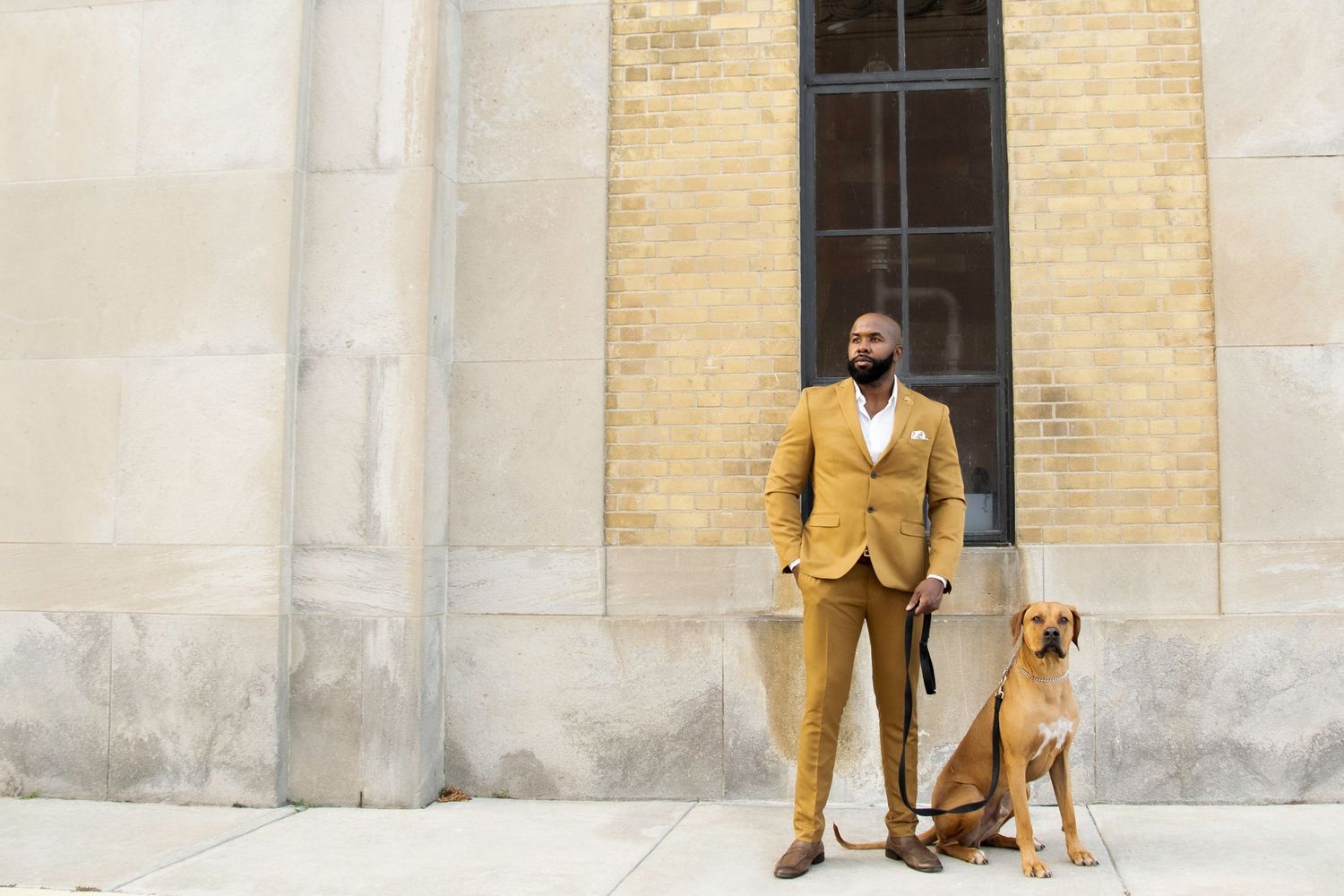 A well-dressed Black man in a mustard yellow suit stands confidently beside his Rhodesian Ridgeback dog outside a stone building.