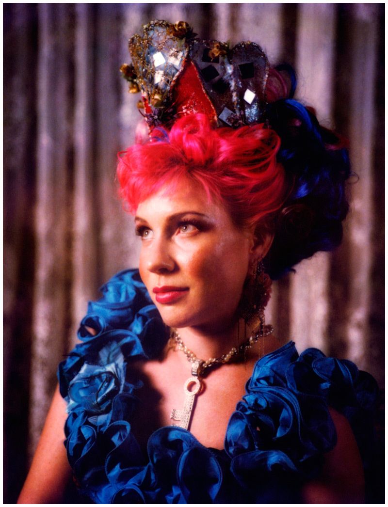 A figure in dramatic lighting wears a blue ruffled collar and red hair with dark floral crown in a theatrical portrait series.