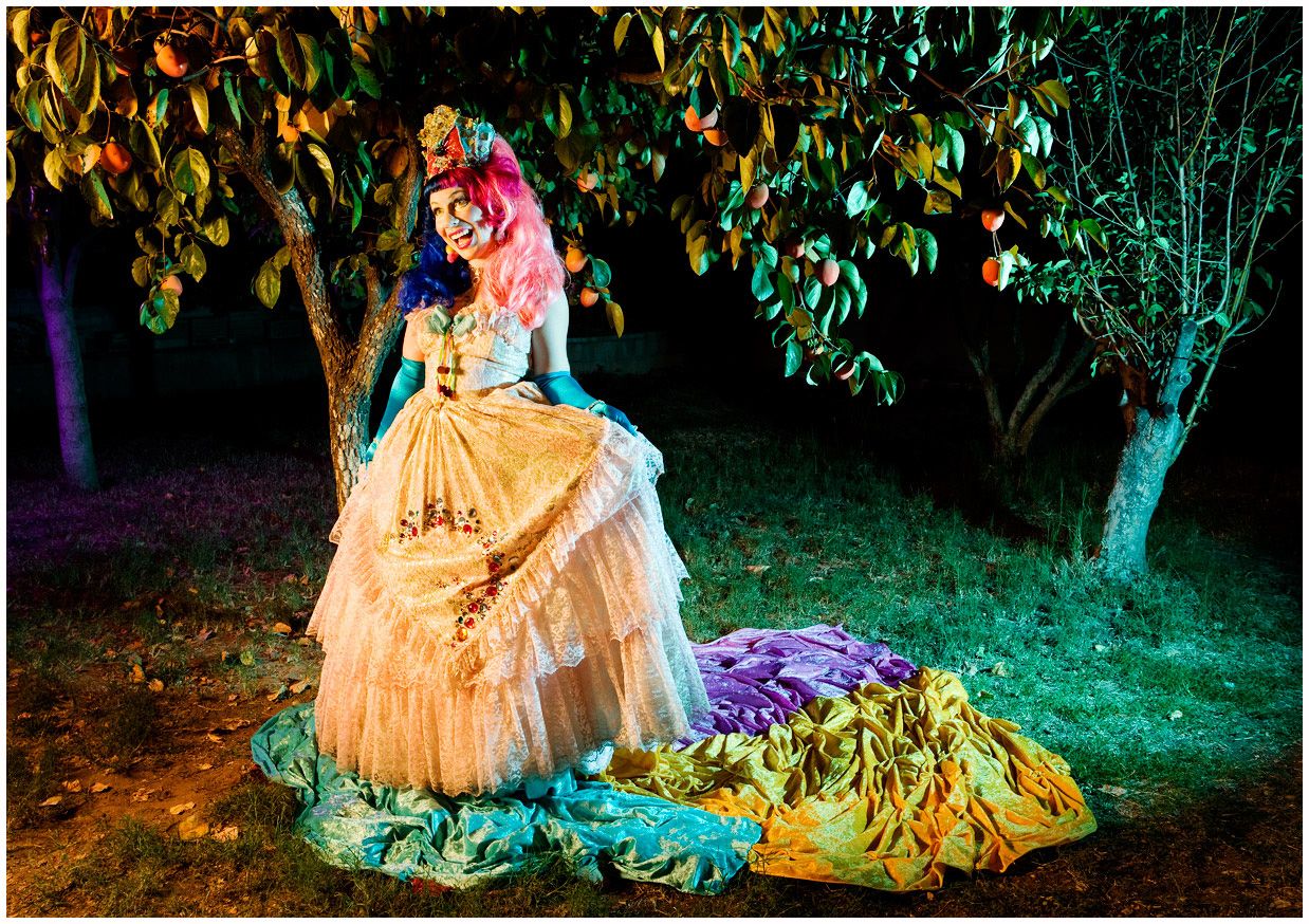 A figure in an elegant gold dress poses near illuminated trees in a dramatic nighttime garden setting.