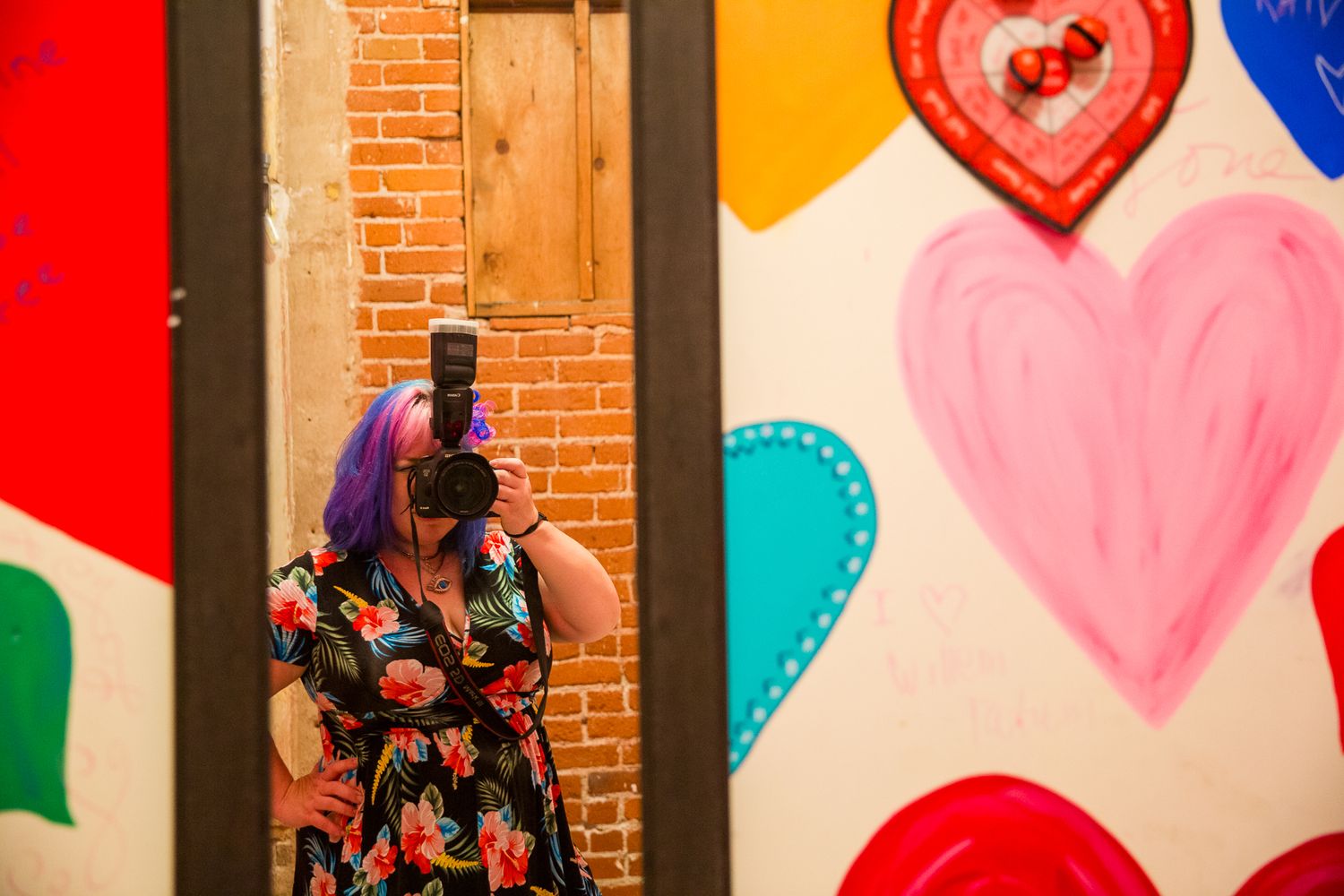 A photographer with blue and pink hair holding a camera looking in a mirror in a bathroom with rainbow hearts on the wall