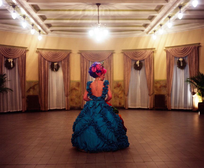 A figure in a dramatic teal ballgown and pink headpiece stands alone in an elegant ballroom with draped windows.