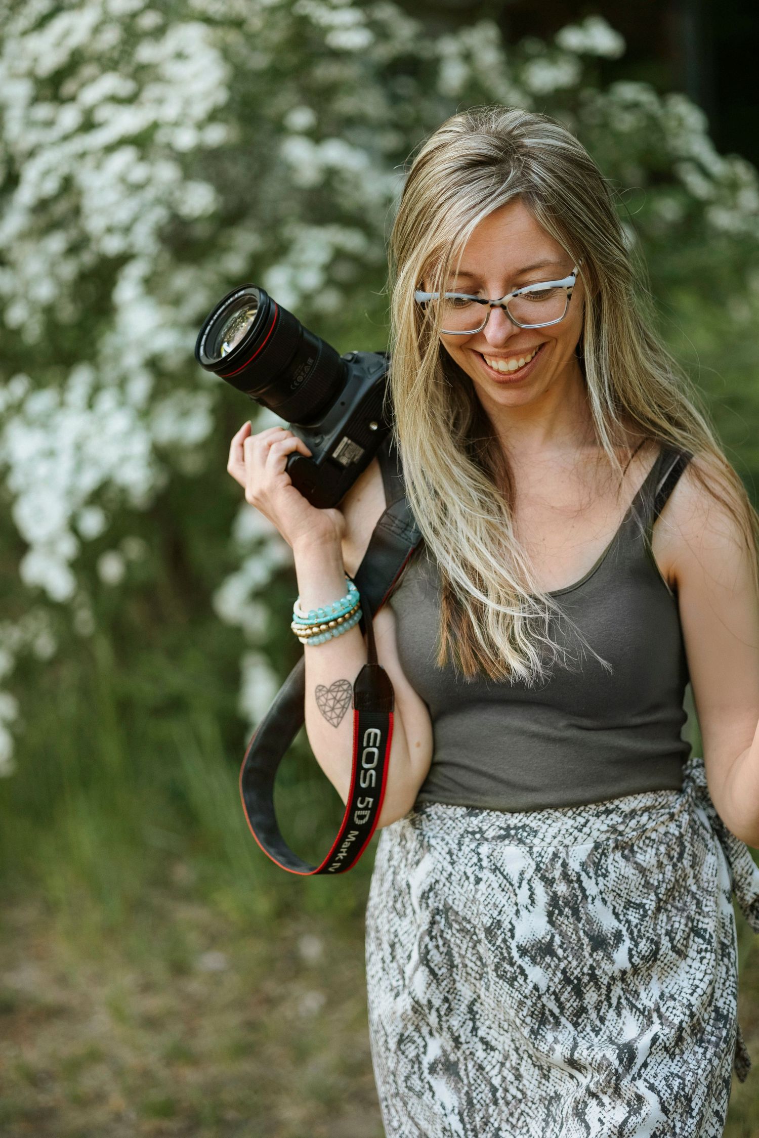 Smiling blonde woman with glasses holding a Canon EOS DSLR camera outdoors near white flowering trees.
