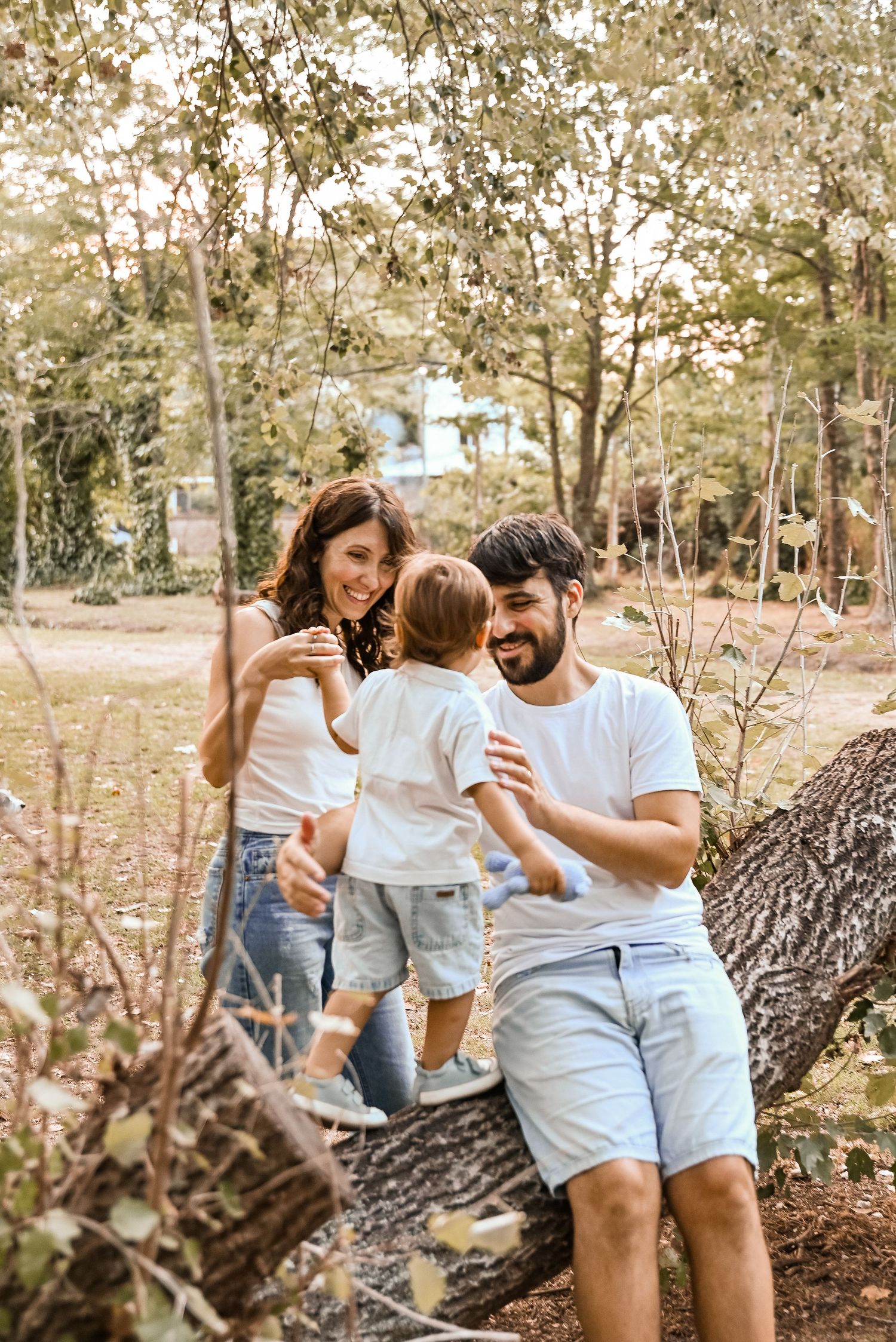 Happy young family with toddler enjoying a sunny day outdoors, sitting on a fallen tree in a lush green park.
