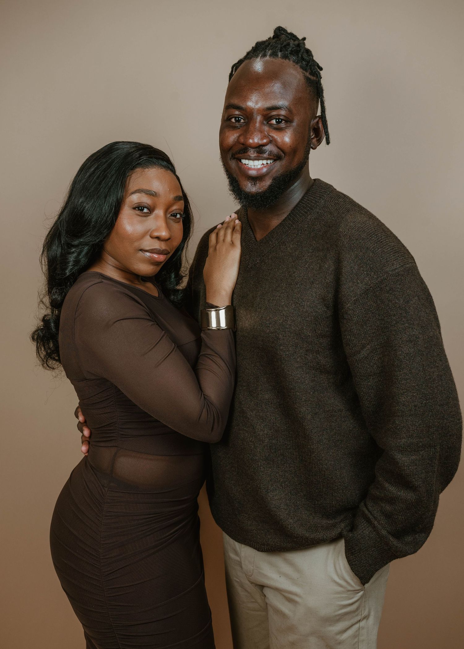 A stylish couple poses together in coordinating brown outfits against a neutral beige background in a professional portrait.