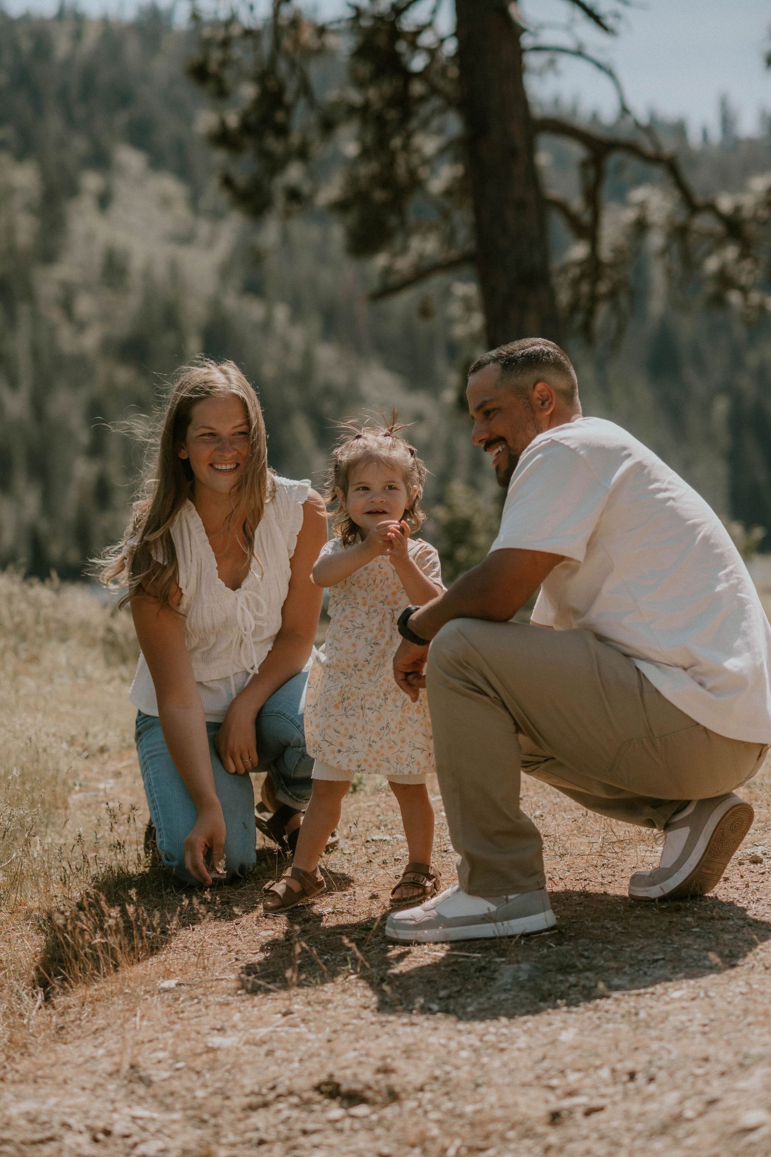 A happy family with a young toddler girl poses outdoors on a sunny mountain trail surrounded by pine trees.