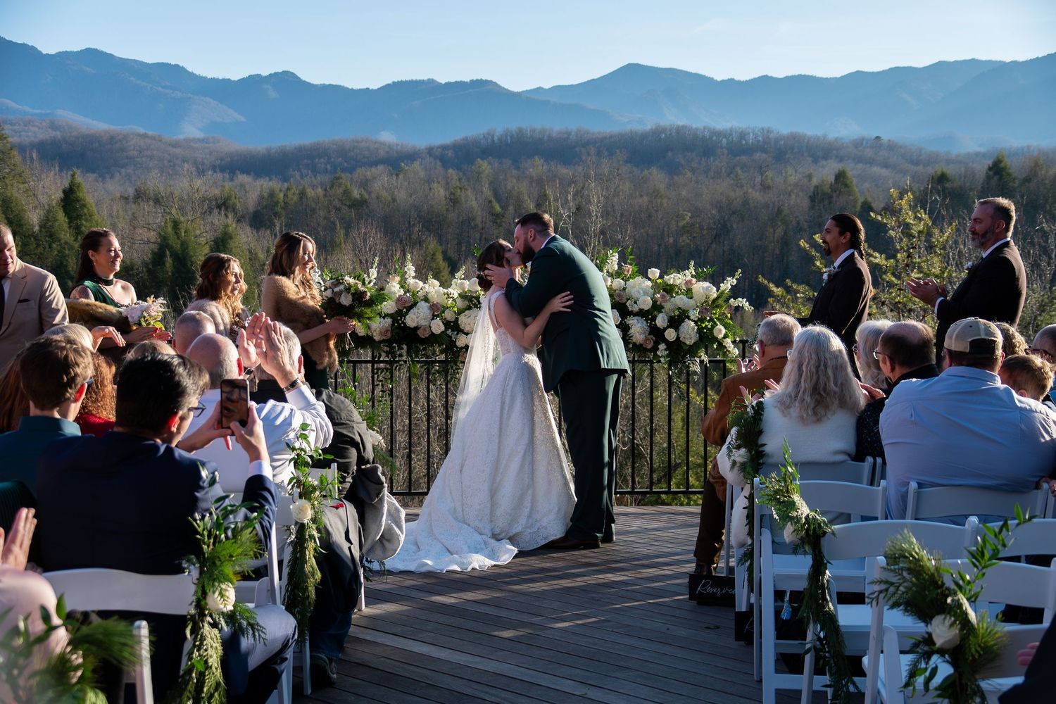 Couple celebrating at outdoor wedding with rainbow in sky, man pointing up, woman in white dress, parking lot setting.