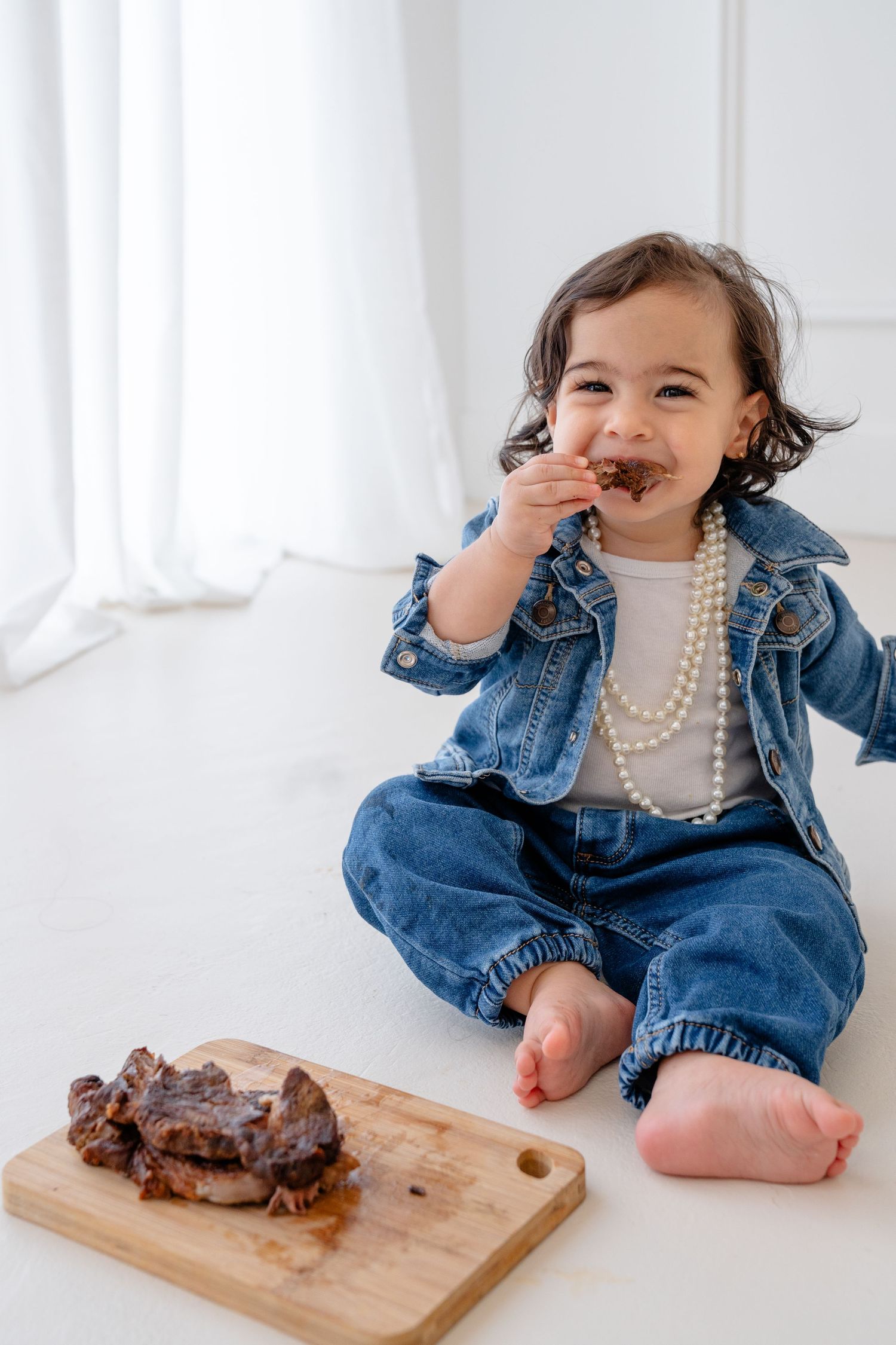 Adorable toddler in denim outfit and pearl necklace sitting on floor eating steak from a wooden cutting board.