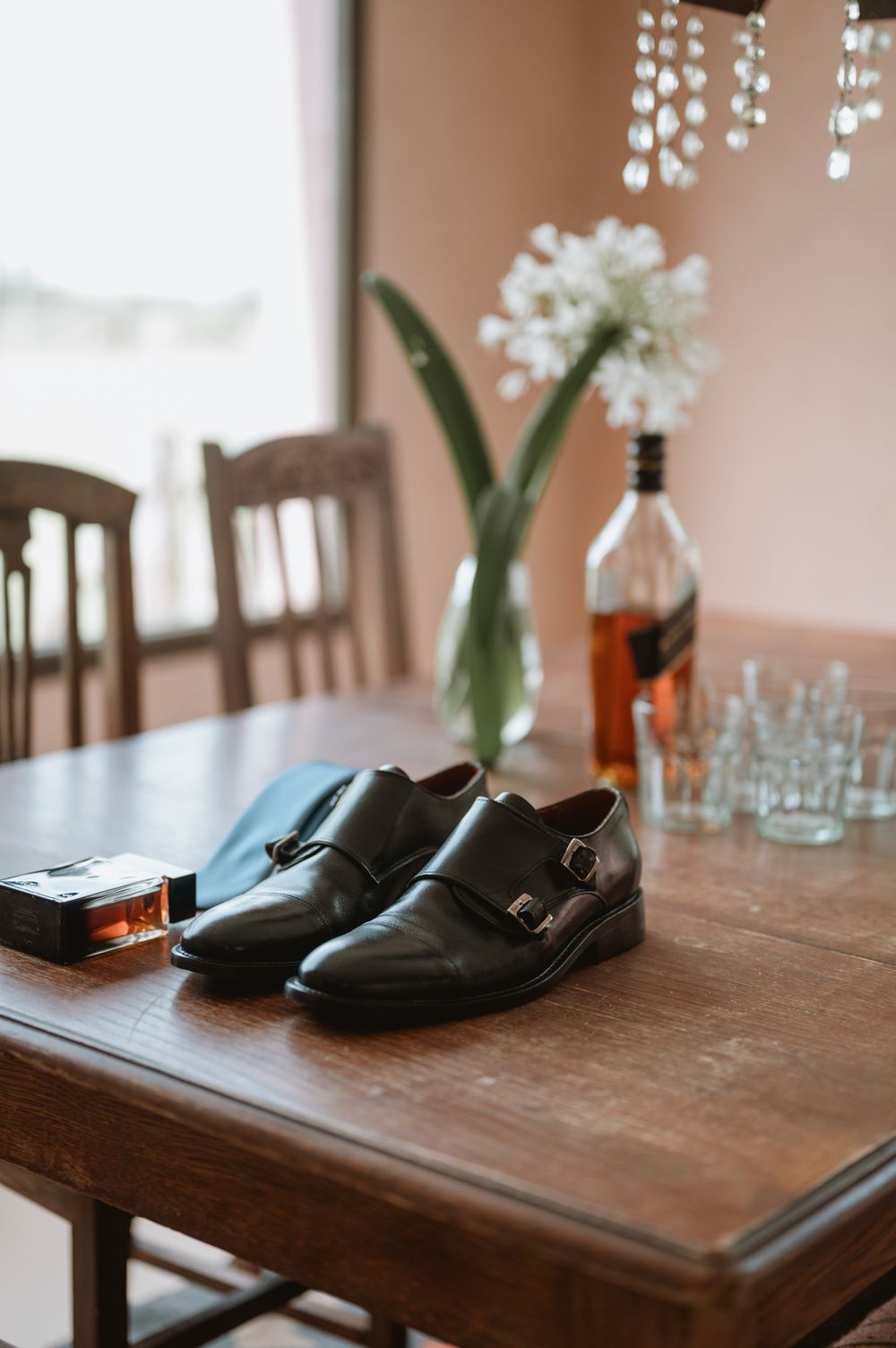 Black monk strap dress shoes on a wooden table with whiskey bottle and glasses.