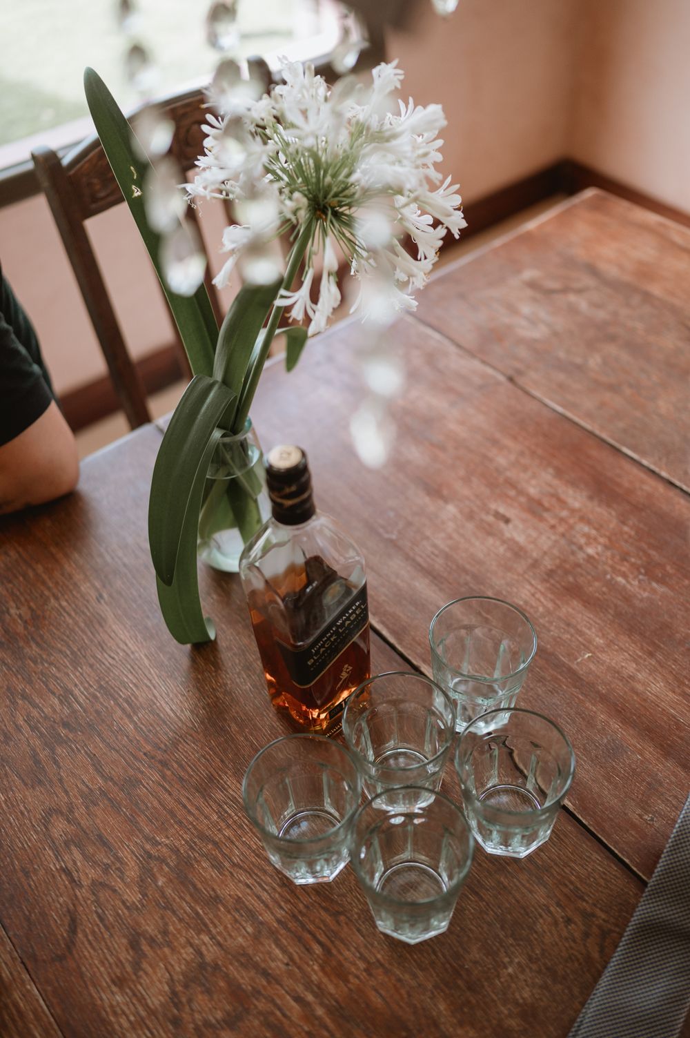 Whiskey bottle with empty glasses and white flowers in a vase on a wooden table.