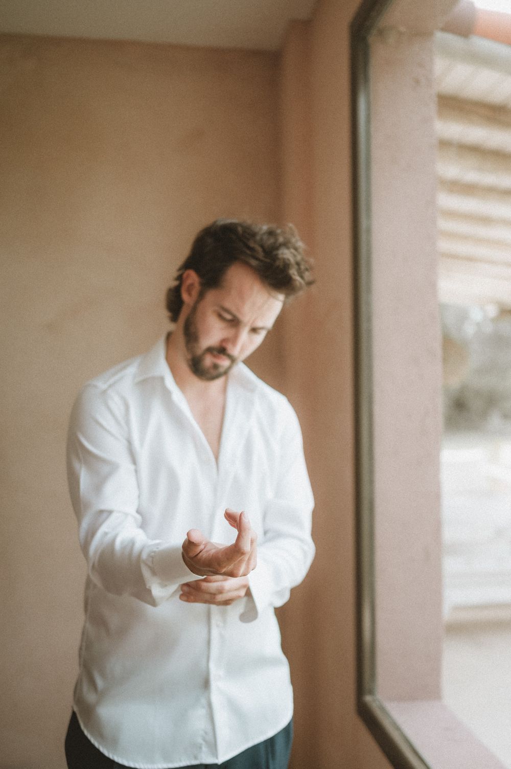Groom in a white shirt adjusting his cufflinks while standing near a window indoors.