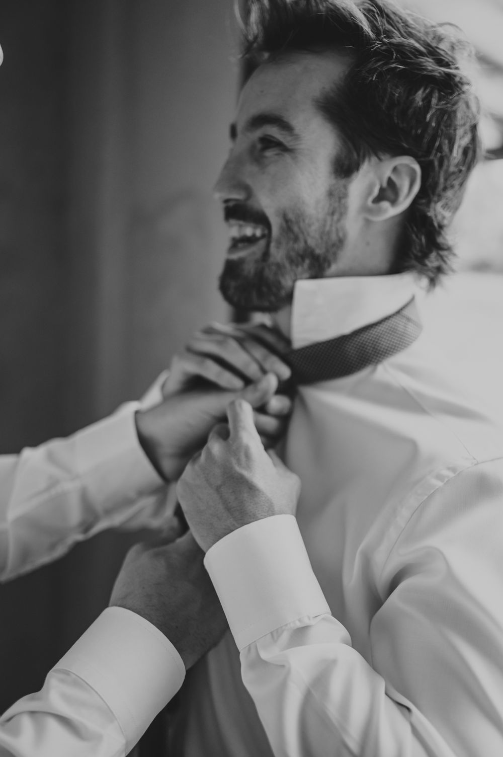 Smiling groom having his tie adjusted by a helper in a black and white wedding photo.