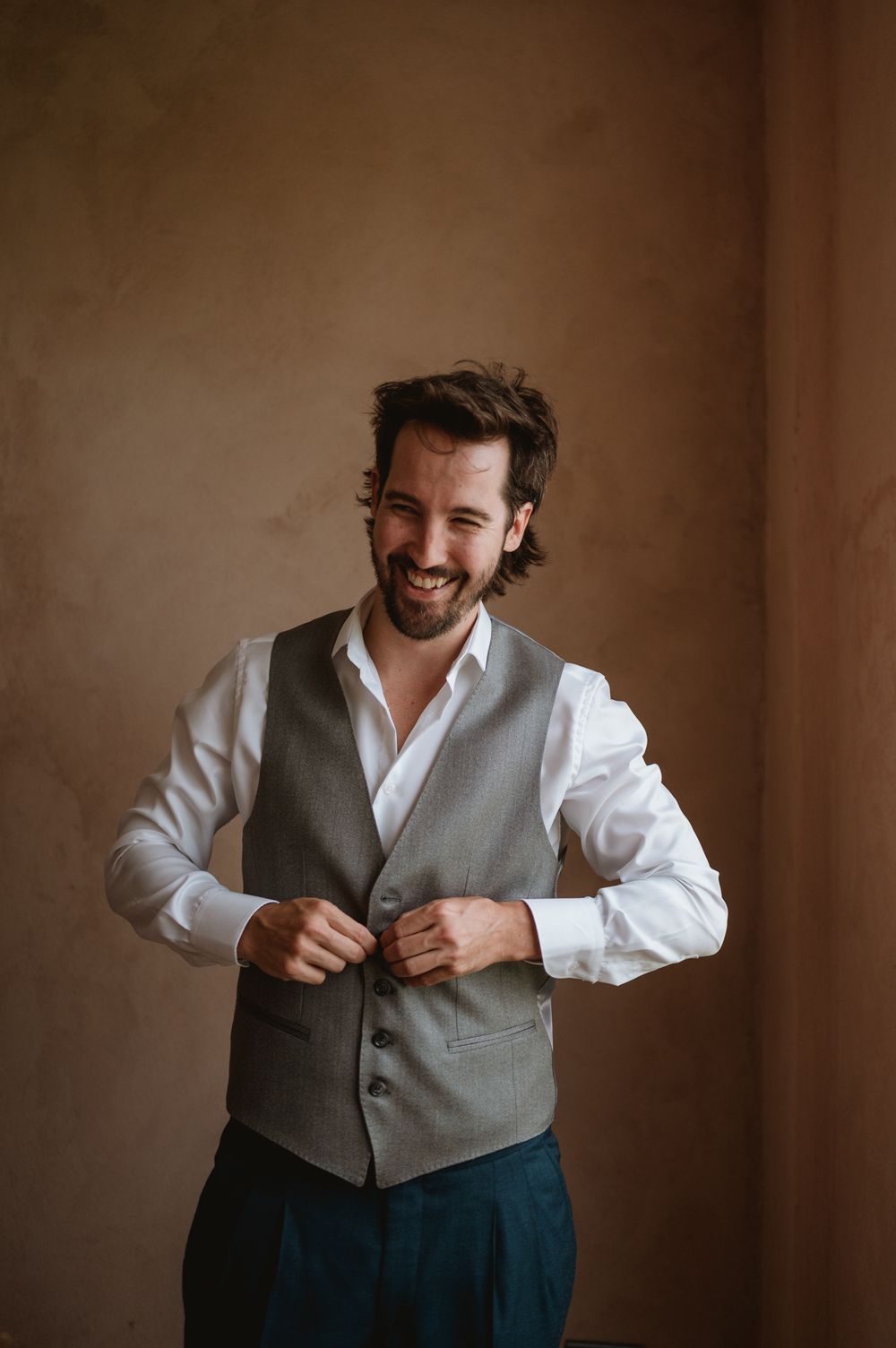 Happy groom buttoning his grey vest over a white shirt before his wedding ceremony.