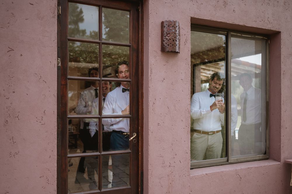 Groomsmen reflected in windows of a pink stucco building during wedding preparations.