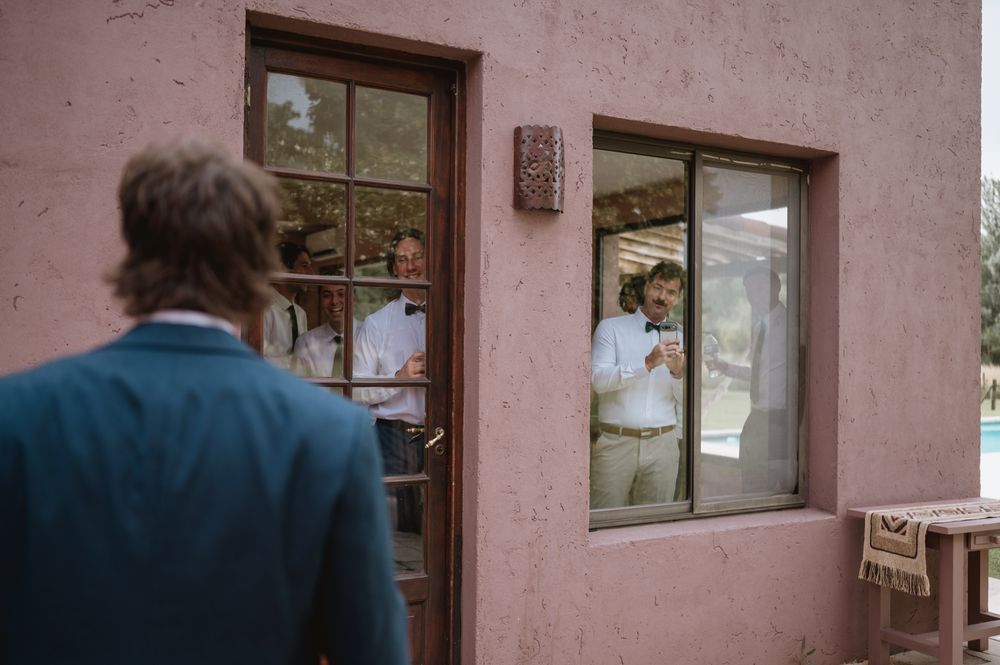 Groomsmen visible through windows of a rustic pink building on a wedding day.